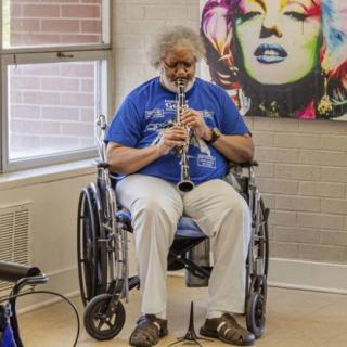A person in a wheelchair plays the clarinet indoors beside colorful wall art.