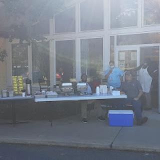 An outdoor setup featuring a food and beverage service table, with people preparing to serve.