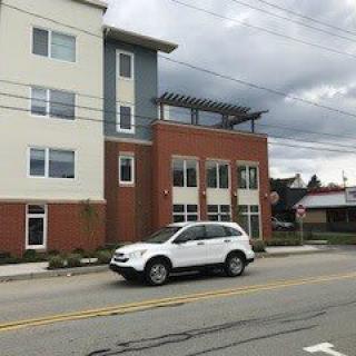 A white car is parked on a street in front of a modern multi-story building under an overcast sky.