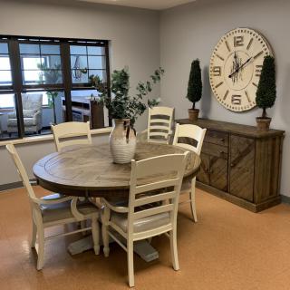 A cozy dining room with a round table and chairs, large clock and plants.