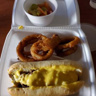 A meal consisting of a cheese-covered hot dog, onion rings, and a side dish served in a disposable container.
