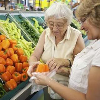 An older woman and a younger woman examining an orange bell pepper while shopping in a grocery store filled with fresh produce.