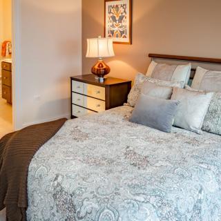 A cozy bedroom with a neatly made bed featuring multiple pillows, a patterned comforter, and a nightstand with a lamp.