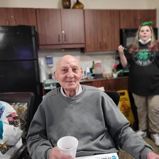 An elderly man smiling while holding a cup, seated in a kitchen setting with a caregiver standing nearby.