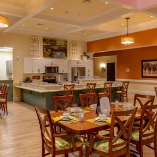 A warmly lit restaurant dining area featuring neatly arranged tables and chairs, with an open kitchen in the background.