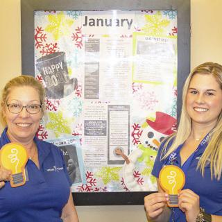 Two women smiling while holding awards in front of a decorated bulletin board.