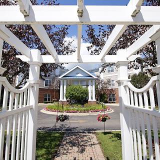 A scenic view of a white pergola leading to a building surrounded by trees and landscaping.