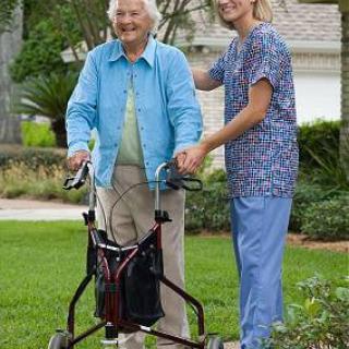 An elderly woman with a walker is assisted by a caretaker in an outdoor setting.