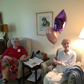 Two elderly women enjoying a moment together with balloons in a cozy indoor setting.