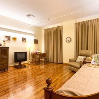 A cozy bedroom with wooden floors and vintage furniture, featuring an armchair, desk, TV, and large curtains.