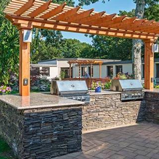 A backyard outdoor kitchen with two grills, a stone countertop, and a wooden pergola.