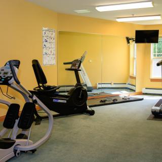 A small gym with exercise equipment including a treadmill, elliptical machine, and a TV on the wall.