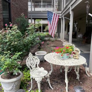 A cozy outdoor patio setup featuring a decorative table and chair, surrounded by potted plants and an American flag.