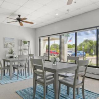 A well-lit dining area with wooden tables and chairs, large windows, and a ceiling fan.