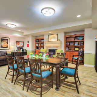 A cozy dining area featuring a central table surrounded by chairs, with bookshelves, artwork, and a fireplace enhancing the ambiance.