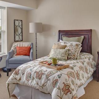 A cozy bedroom with a single bed featuring floral bedding, an armchair, and a lamp by the window.