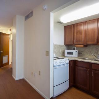 A view of a kitchen area with wooden cabinets, a stove, and a microwave, adjacent to a hallway in an apartment.