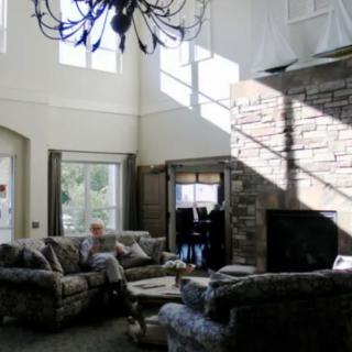 A spacious living room with high ceilings, featuring a chandelier, large windows, a stone fireplace, and comfortable sofas.