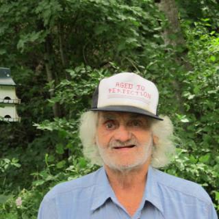A man wearing a hat stands outdoors with trees in the background.