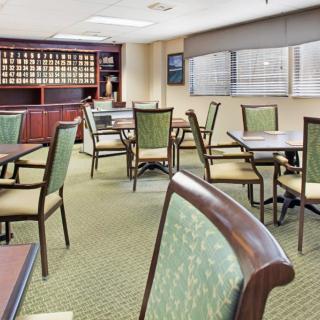A well-lit dining area with tables and chairs, carpeted flooring, and large windows.