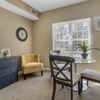 A cozy living room corner with a small round table, chairs, an armchair, and a window providing natural light.