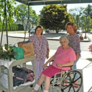 Two caretakers accompany an elderly woman in a wheelchair outside a medical facility.