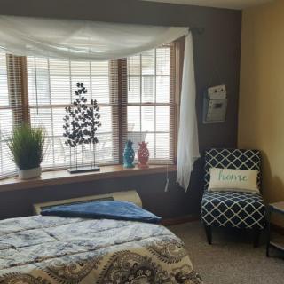 A cozy bedroom with a large bay window, decorated with plants and colorful vases.