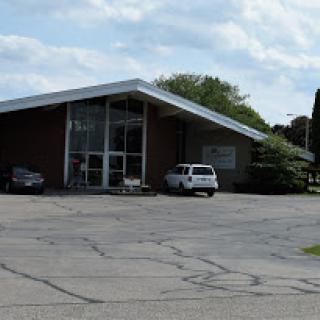 A modern building with a distinctive roof design and parked cars in the lot under a partially cloudy sky.