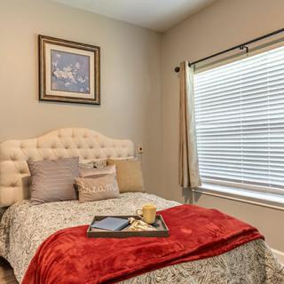 A cozy bedroom with a neatly made bed, featuring decorative pillows and a red throw blanket, complemented by wall art and natural light from the window.