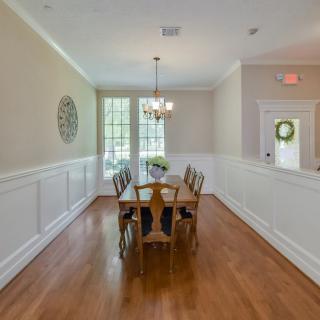 A bright dining room with a wooden table and chairs, featuring a chandelier and large windows.
