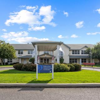 A two-story residential building with a welcoming entrance, surrounded by well-maintained greenery and set against a bright blue sky with scattered clouds.