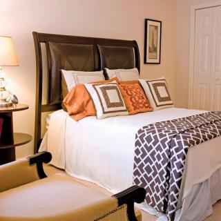 A cozy bedroom with a neatly made bed featuring decorative pillows and a patterned blanket, complemented by warm lighting from a lamp.