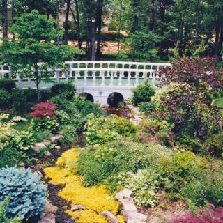 A picturesque garden with a small white bridge surrounded by lush greenery and colorful plants.
