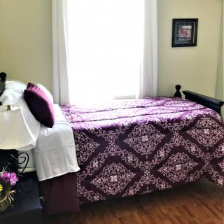 A cozy bedroom with a purple-patterned bedspread and sunlight streaming through the window.