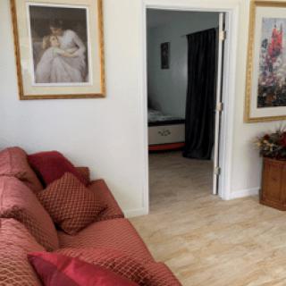 A cozy living room with a red sofa, decorative pillows, and framed paintings on the wall, leading to a bedroom through an open doorway.
