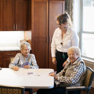 A caregiver interacts with two elderly women at a communal table, highlighting the importance of companionship and care.