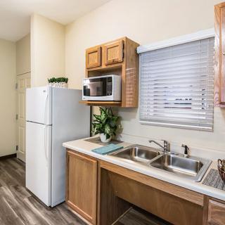 A compact kitchen with essential appliances, featuring a refrigerator, microwave, dual sink, and wooden cabinets.