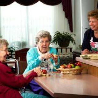 Three elderly women enjoying a conversation while preparing apples in the kitchen.