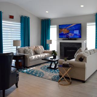 A modern living room with neutral-colored sofas, a wall-mounted television above a fireplace, and vibrant teal curtains.