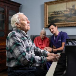 An elderly man plays the piano while two people, possibly a caregiver and a fellow resident, watch and enjoy in a nursing home setting.