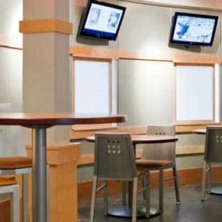 A modern restaurant interior with tables and chairs, featuring wall-mounted TV screens.