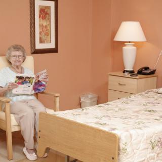 An elderly woman sits in a comfortable chair reading in a cozy bedroom.