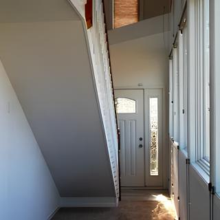A narrow hallway with a staircase above and a front door at the end, illuminated by natural light through side windows.
