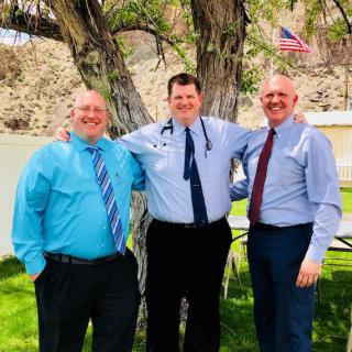 Three men in formal attire smiling outdoors beside a tree.
