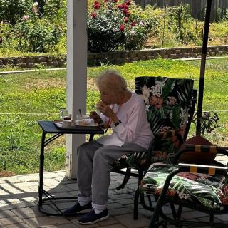 An elderly person enjoys a meal while sitting on a patio, surrounded by nature.