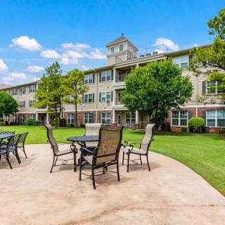 A scenic view of a building with a patio featuring tables and chairs, surrounded by a well-maintained lawn and trees under a clear blue sky.