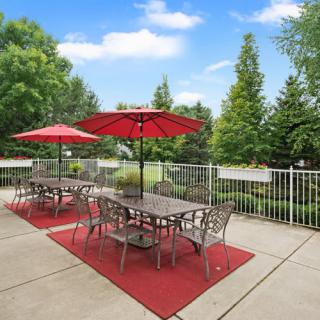 An outdoor patio area with tables and chairs under red umbrellas, surrounded by a metal fence and lush greenery.