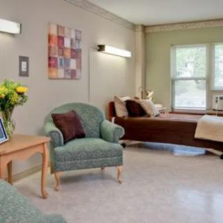 A cozy hospital room with a bed, chairs, and a table with flowers under soft lighting.