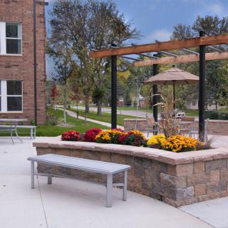 A serene outdoor patio area with a bench, flower bed, and pergola near a brick building.
