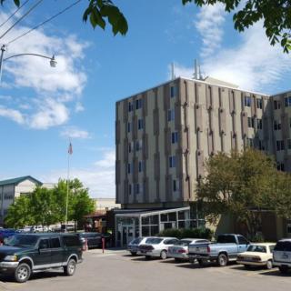 A street view featuring a multi-story building surrounded by parked cars and trees under a partly cloudy sky.
